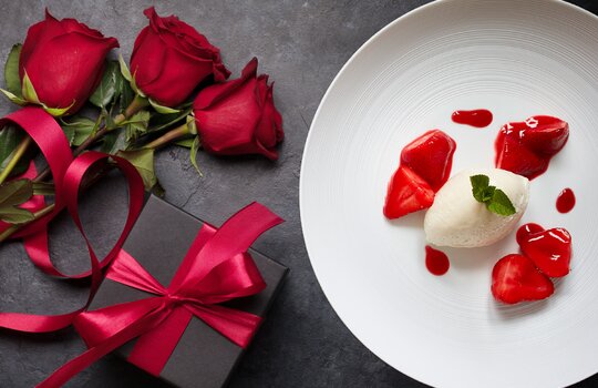 Ein Dessert, drei rote Rosen und ein Geschenk auf schwarzem Hintergrund