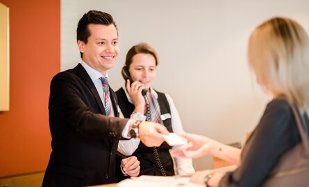 Ein lächelnder Rezeptionsmitarbeiter des Romantik- und Wellnesshotels Deimann überreicht einer Dame ihre Hotelzimmerkarte