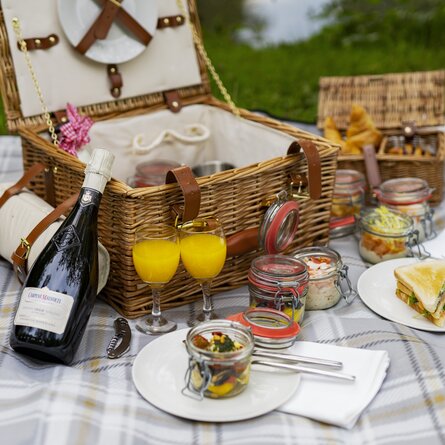 Ein reichhaltig gefüllter Picknickkorb auf einer karierten Decke mit Sekt, Orangensaft und Dips in kleinen Gläsern 