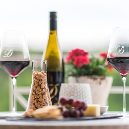 Zwei Gläser Rotwein, eine Flasche Rotwein, Erdnüsse im Glas, shwarze Weintrauben und einen Blumentopf auf dem Tisch.