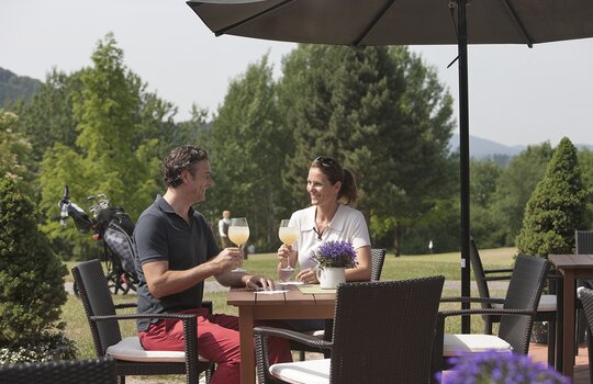 Ein glückliches Ehepaar sitzt auf der Panoramaterrasse des Golf-Cafés ihres Golfhotels in NRW und trinkt Cocktails