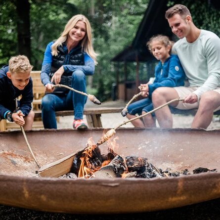 Eine vierköpfige Familie sitzt in der Natur um eine Feuerstelle in ihrem Familienurlaub Sauerland