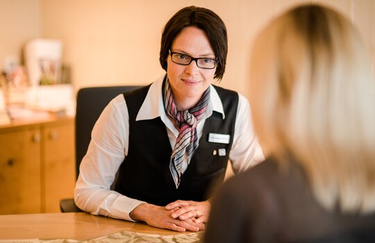 Eine Rezeptionistin des Romantik- und Wellnesshotels Deimann hört einer Dame zu und lächelt sie an 