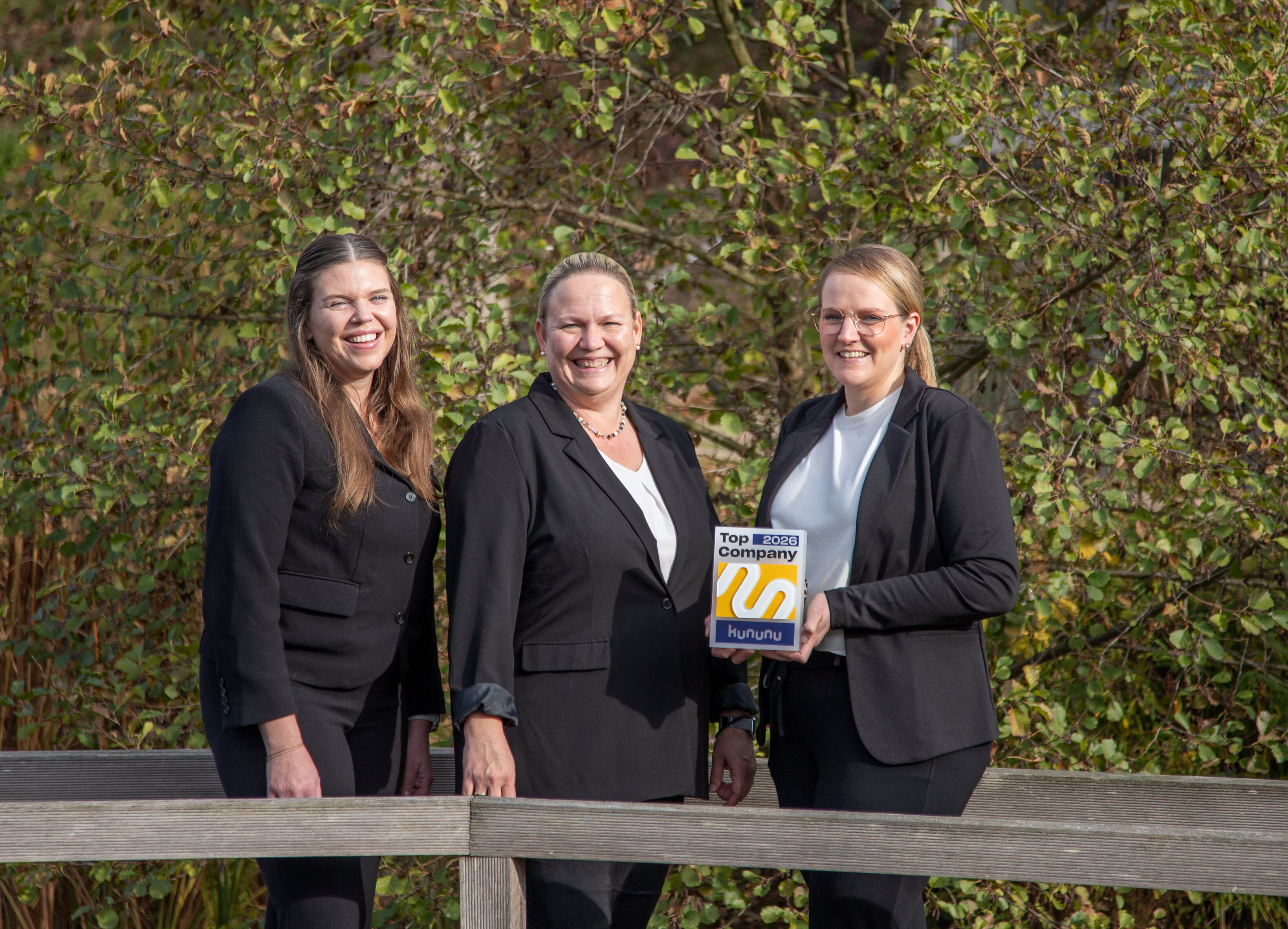Drei Frauen in schwarzen Blazern stehen draußen vor einer grünen Hecke; eine hält eine Auszeichnung mit der Aufschrift 'Top Company 2026 Kununu'.