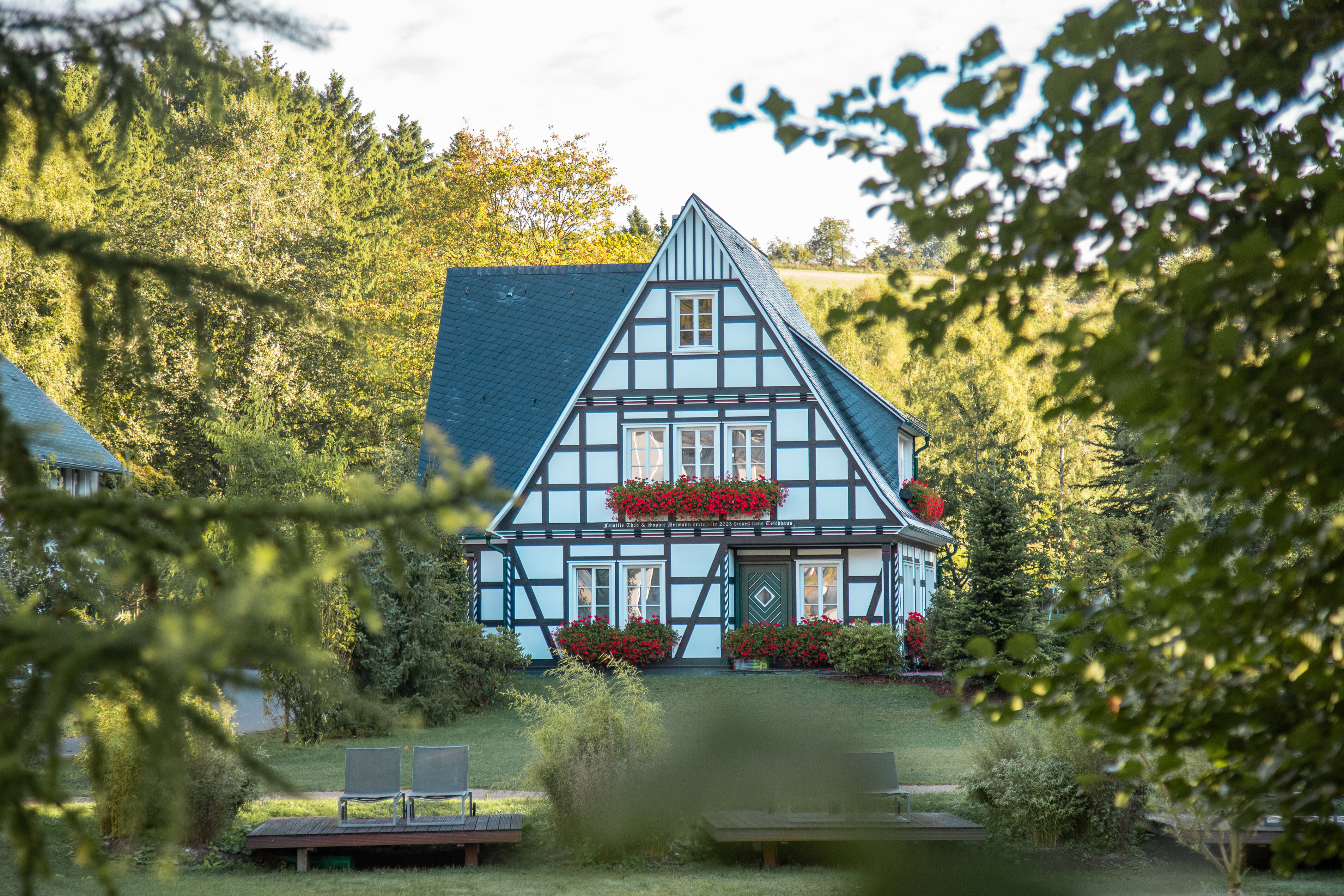 Historisches Fachwerkhaus mit blauem Dach, weißen Infill und rotem Blumenschmuck in Fensterkästen, umgeben von Gartenbäumen; zwei Bänke im Vordergrund.