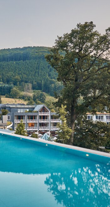 Außenansicht eines Hotels mit Infinity-Pool auf der Dachterrasse, Blick über Gartenanlage und Waldhügel; zentraler Baum; mehrstöckige Hotelgebäude im Hintergrund; Spiegelungen im Wasser.