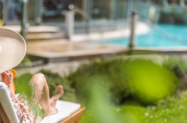 Frau mit Sonnenhut sitzt auf einer Liege am Pool