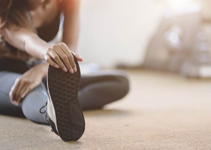 Close-up of a person seated on a gym floor, holding the sole of a sneaker with one hand while extending a leg, with gym equipment blurred in the background.